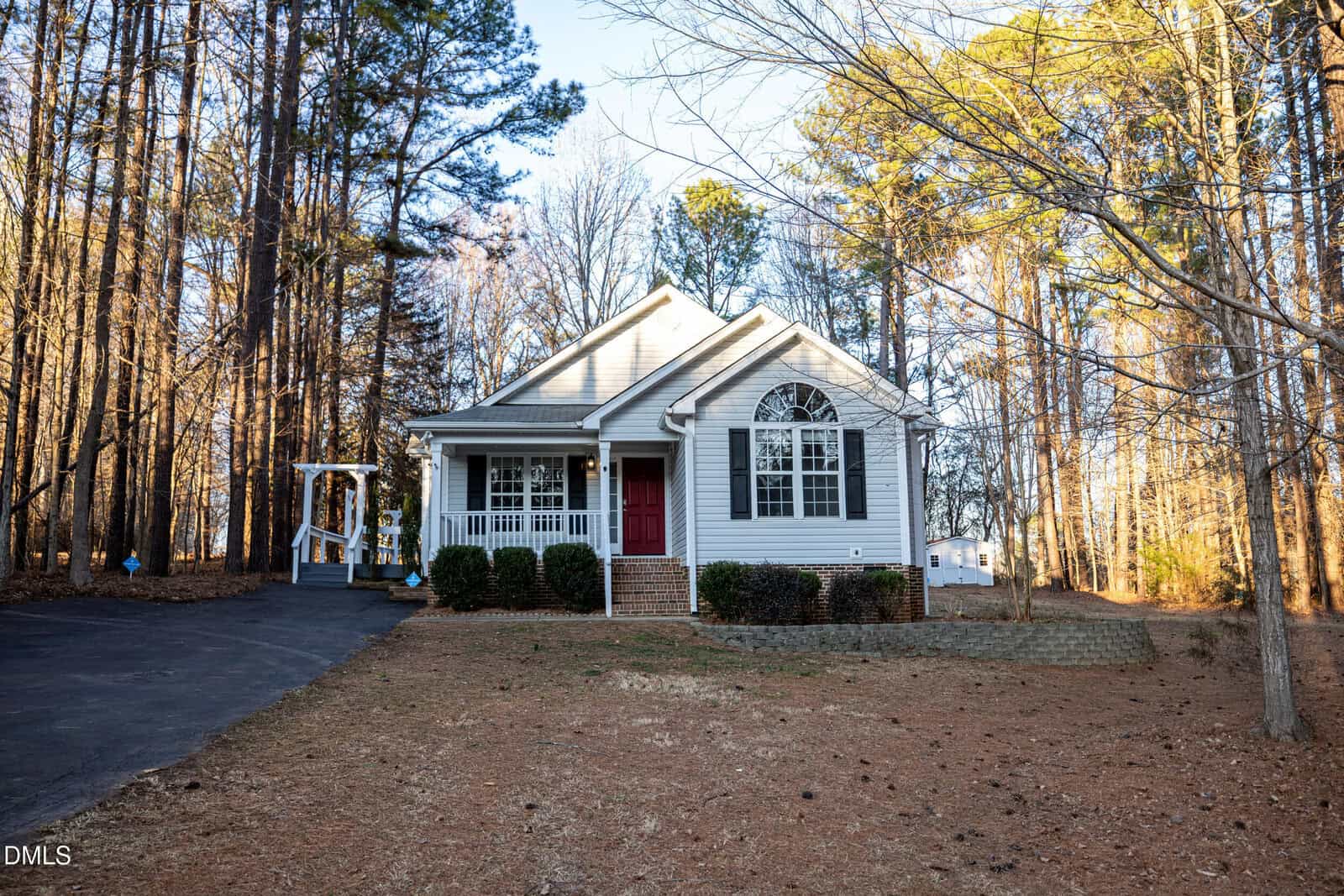 A small, single-story white house with a red front door and arched window sits on 50 Millstone Drive in Franklinton, surrounded by tall trees and a large, partially wooded yard. A paved driveway leads up to the inviting front porch.