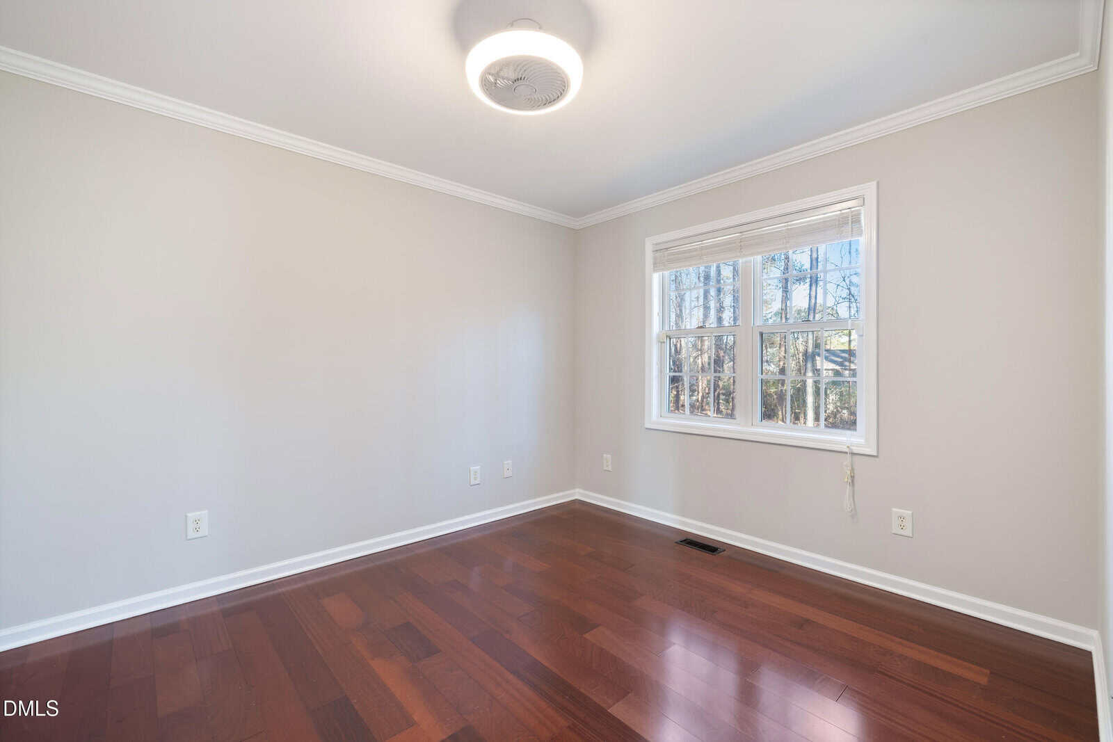 Empty room with beige walls, large window with white trim, dark wood floor, ceiling light, and some visible electrical outlets. Sunlight streams in through the window, revealing trees outside.