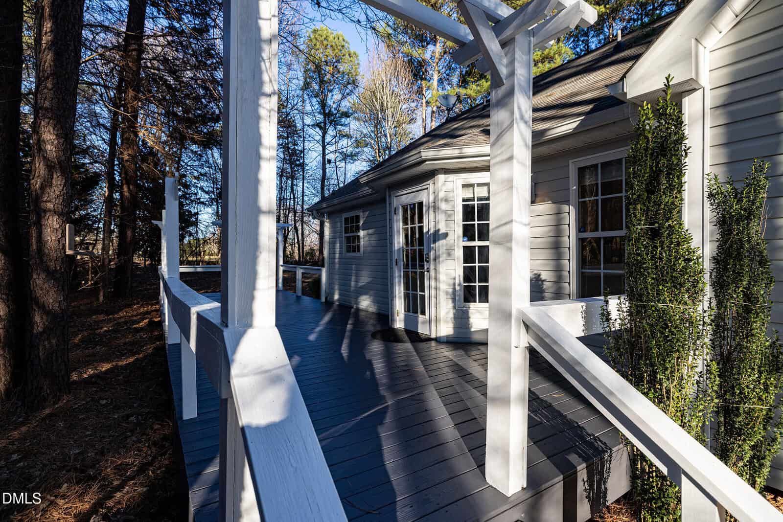 A white wooden deck with railings wraps around a light-colored house, surrounded by tall trees. Sunlight creates sharp shadows across the deck and house exterior.