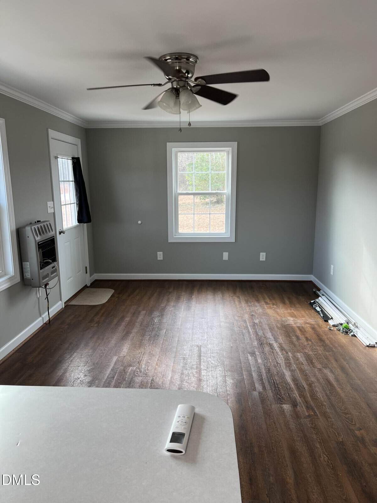 A mostly empty room with hardwood floors, gray walls, a white ceiling, ceiling fan, two windows, a white door, wall heater, and some items near the baseboard; a white countertop and remote are in the foreground.