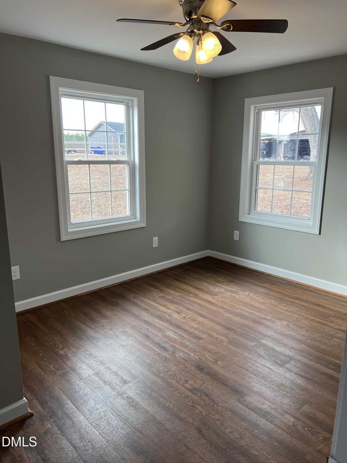 A small, empty room with gray-green walls, two large windows, dark wood flooring, and a ceiling fan with lights. Natural light is coming through the windows, showing an outdoor yard and trees.