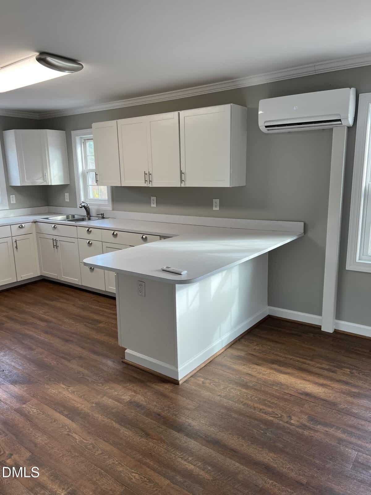 Bright kitchen with white cabinets, light countertops, wood flooring, and a wall-mounted air conditioning unit. A peninsula extends from the counter, and natural light streams in through two windows.