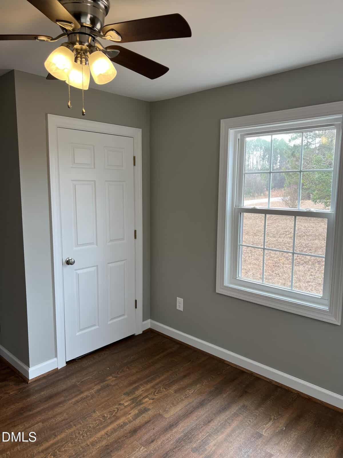 A small empty room with gray walls, a white door, a window with an outdoor view, a ceiling fan with lights, and dark wood flooring.