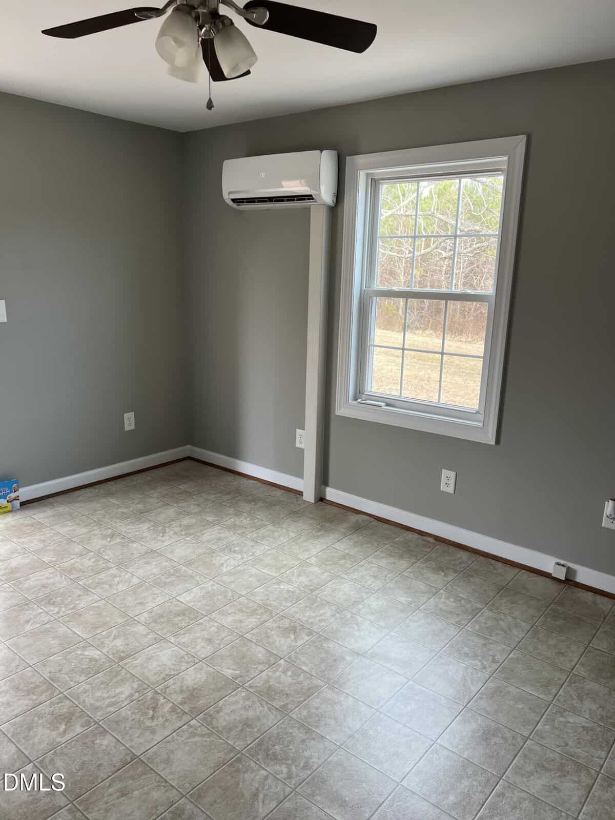 A small, empty room with gray walls, a ceiling fan, tile flooring, a window with white trim, and a wall-mounted air conditioner above the window. There are a few toys in one corner.