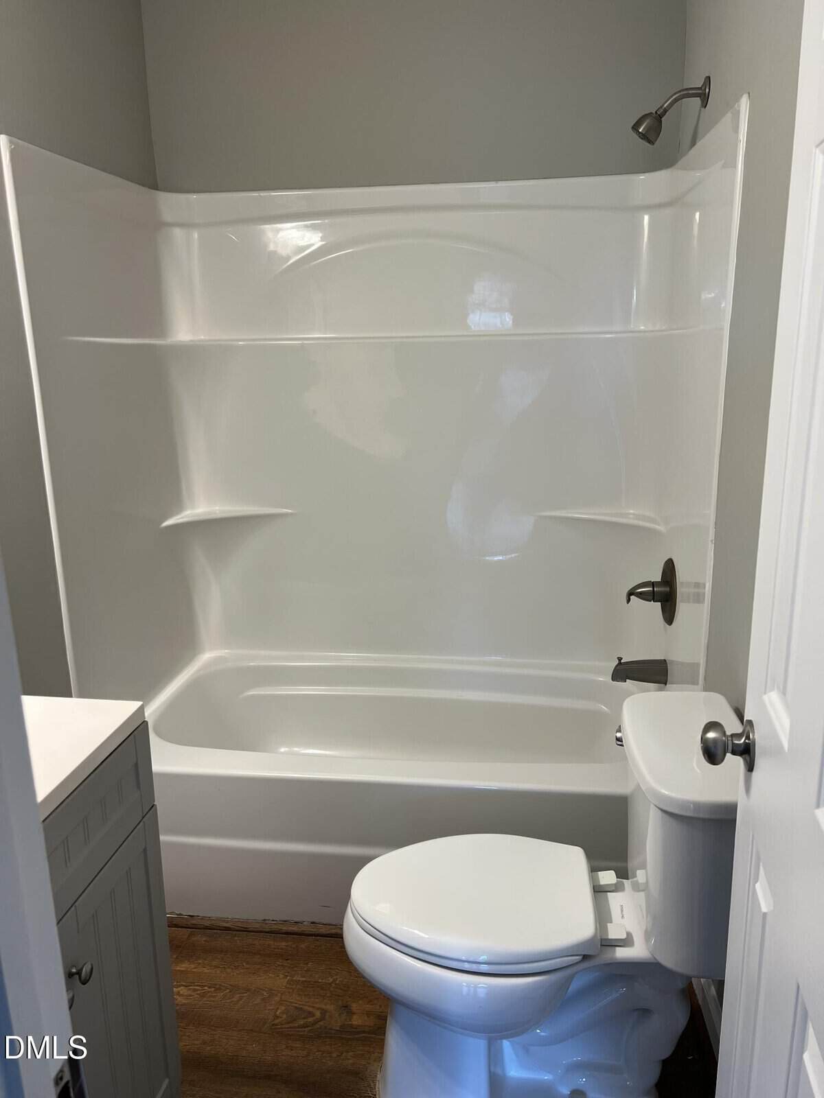 White bathroom with a bathtub-shower combo, white toilet, light gray walls, and wooden floor. A small white vanity is partially visible on the left. Simple, clean, and modern design.