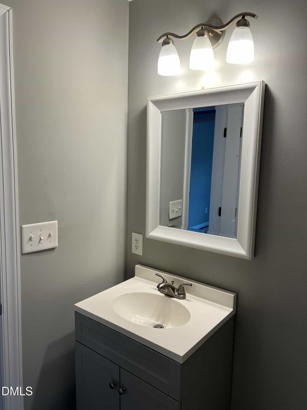 A small bathroom vanity with a white sink, gray cabinet, wall mirror, and three-light fixture above. The wall is painted gray, and there are light switches to the left of the vanity.