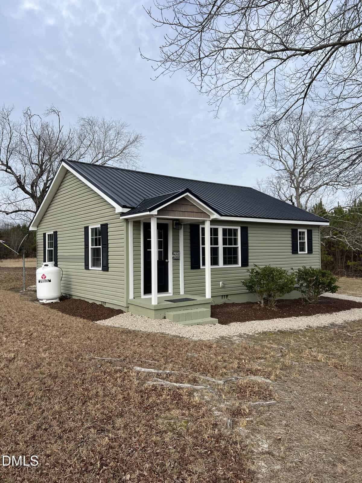 A small, single-story green house with black shutters and a black roof sits on a tidy lot at 317 Brewer Road in Louisburg, featuring a covered front porch, shrubs, propane tank, bare trees, and brown grass.