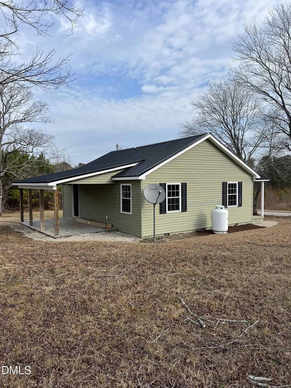 Single-story house with light green siding, dark roof, and black shutters. A covered porch wraps around one side. A satellite dish and white propane tank sit in the yard. Leafless trees and a partly cloudy sky are in the background.