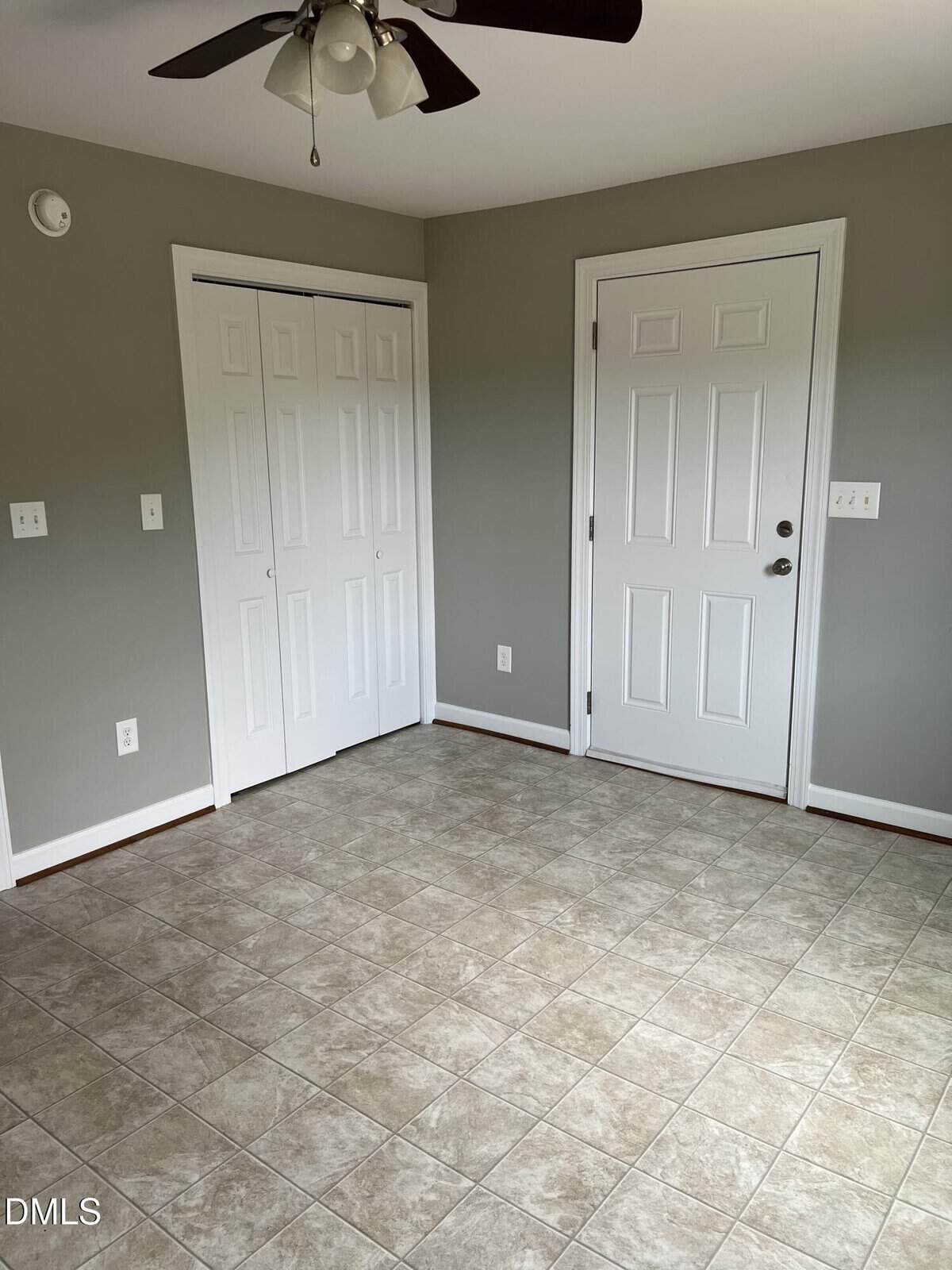 Empty room with beige tiled floor, gray walls, a white door, white double closet doors, a ceiling fan with lights, and a small wall smoke detector.