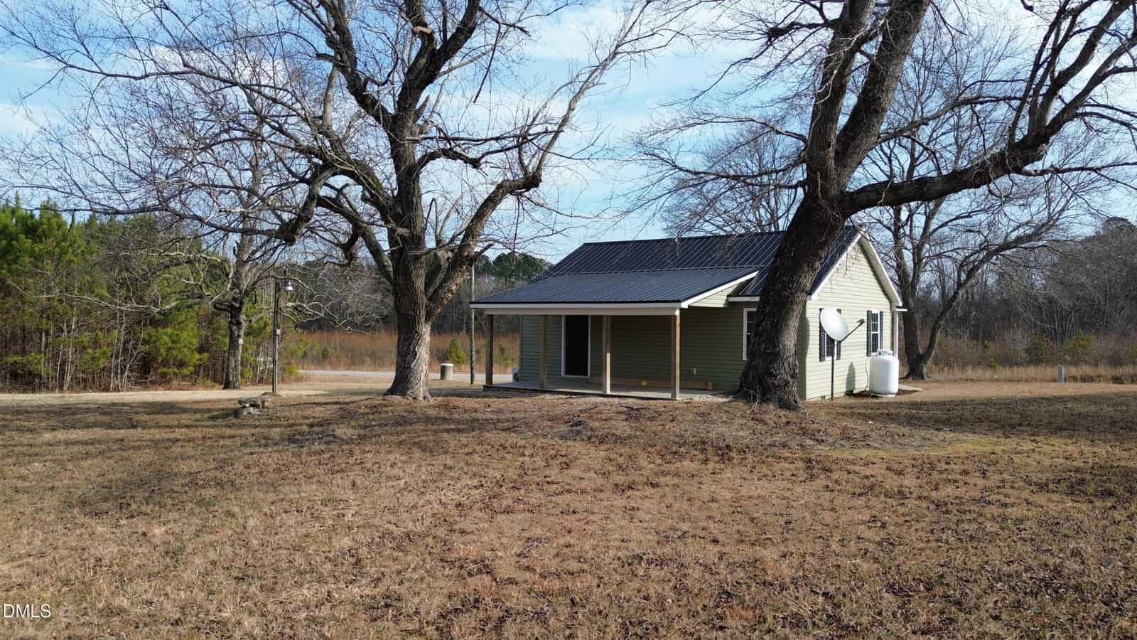 A small, light green house with a black metal roof sits among leafless trees on a dry, grassy yard under a blue sky. A satellite dish is attached to the house, and a propane tank is visible on the right.