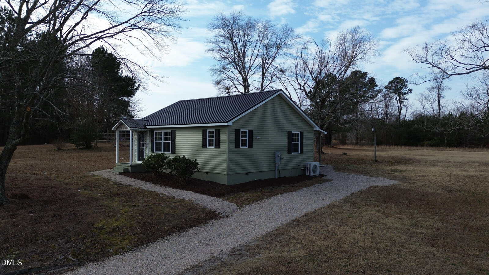 A small light green house with a dark roof and a gravel driveway sits on a large, grassy lot with leafless trees under a partly cloudy sky.
