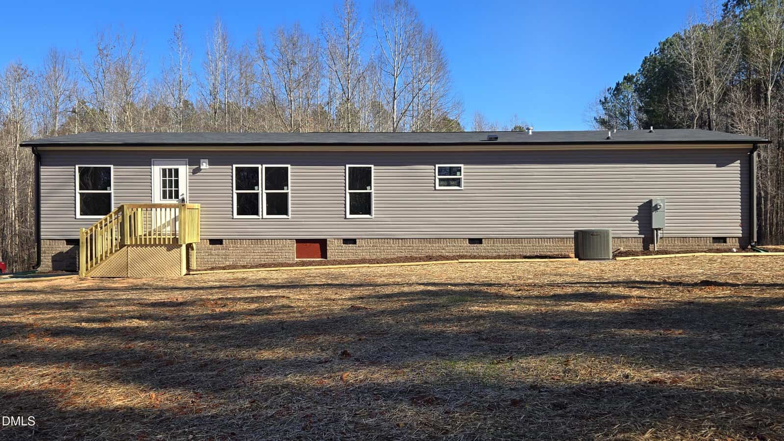 A single-story manufactured home with gray siding, six windows, a small wooden porch with stairs, an outdoor HVAC unit, and a brick foundation, set on a cleared lot bordered by leafless trees.