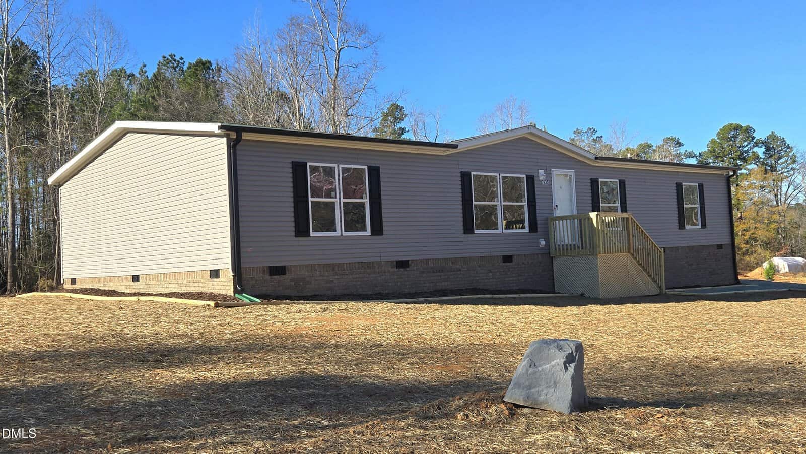 A single-story, gray manufactured home with black shutters and a small front porch sits on a cleared lot at 7635 Sam Hall Road in Oxford, surrounded by bare soil, minimal landscaping, and a backdrop of leafless trees and evergreens.
