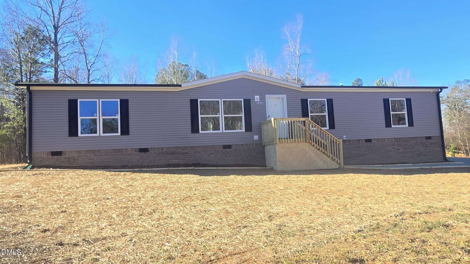 A single-story manufactured home with gray siding, black shutters, and a small wooden front porch, set on a bare lawn with trees and a blue sky in the background.