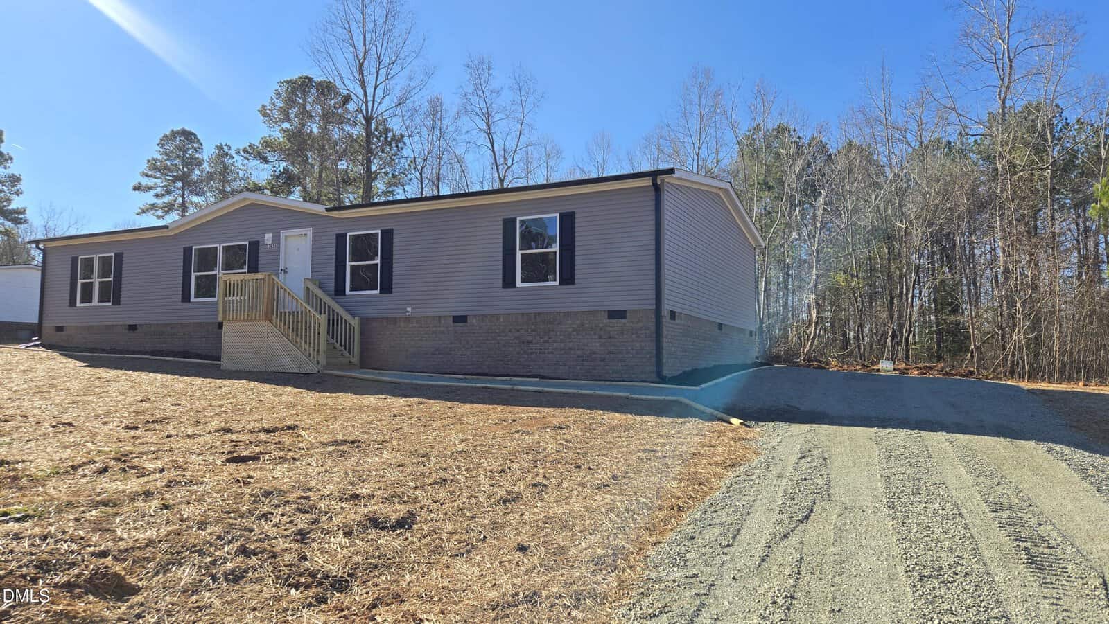 A single-story manufactured home with gray siding, a small covered front porch with railings, brick foundation, and a gravel driveway. The yard is bare with straw and some trees in the background under a clear blue sky.