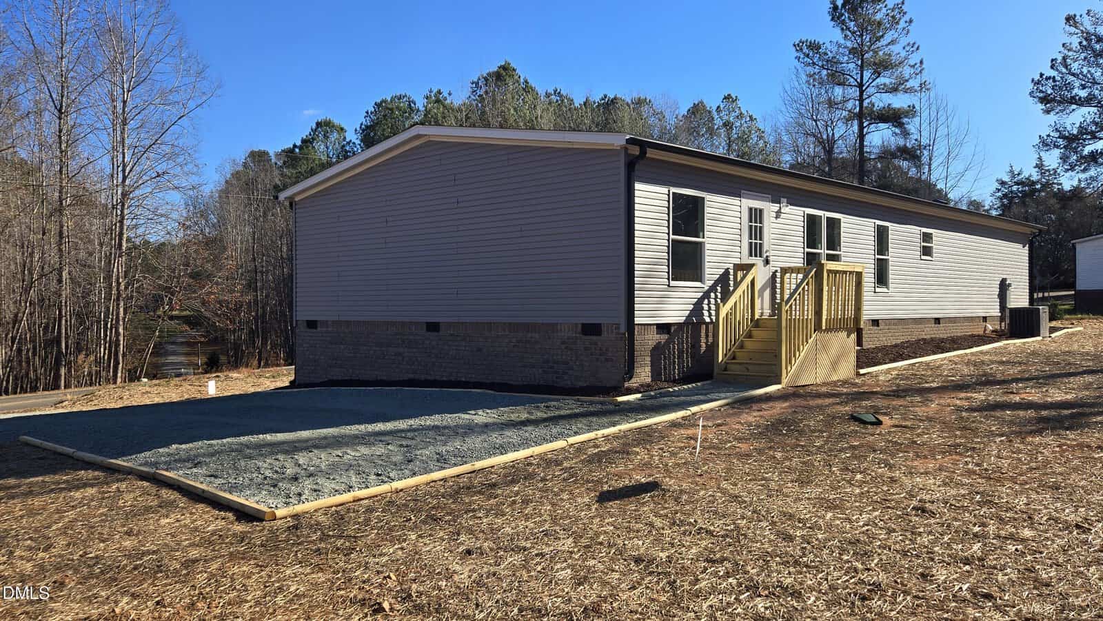 A single-story manufactured home with light-colored siding and a brick foundation, set on cleared land with a gravel area and newly-built wooden steps leading to the main entrance; trees and blue sky in the background.