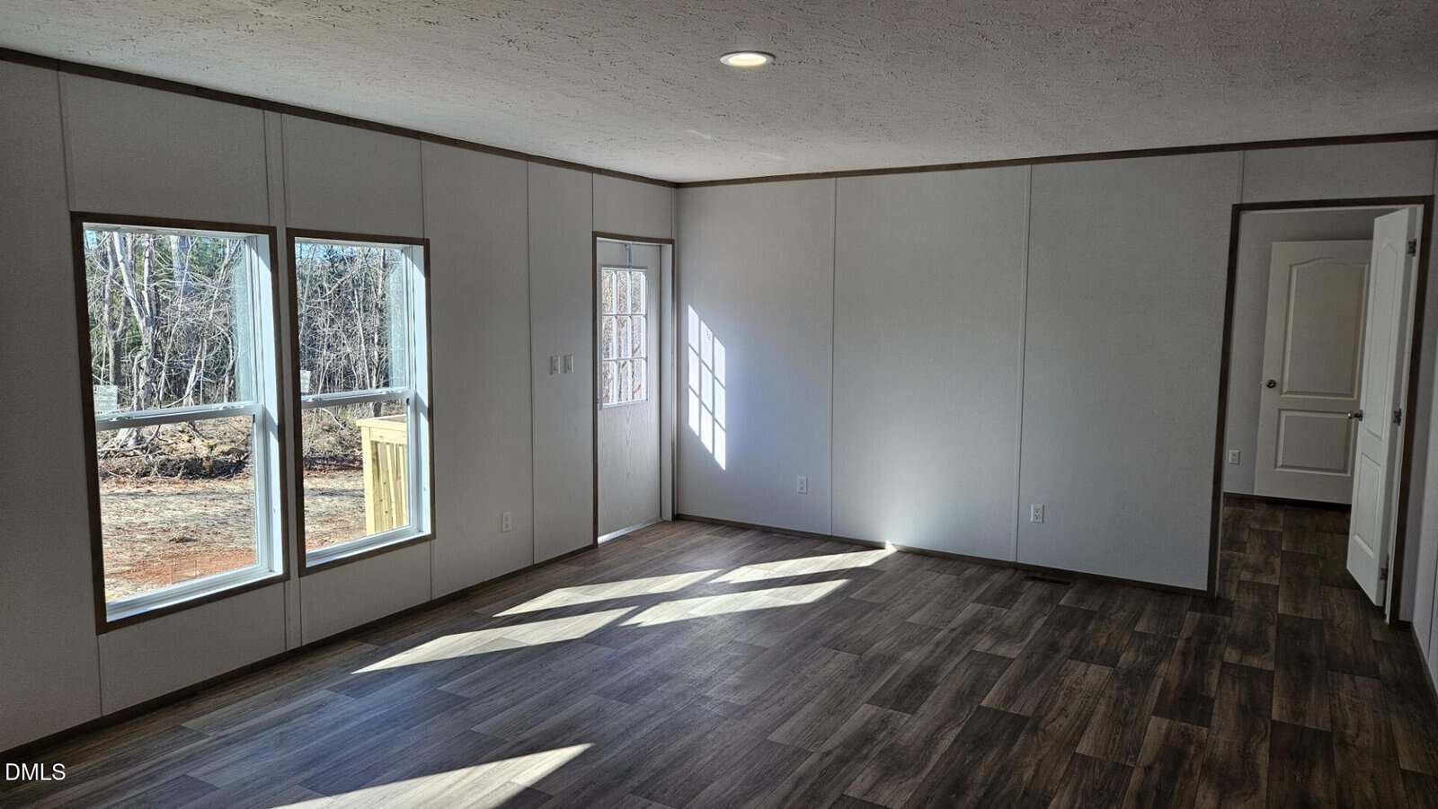 A bright, empty room with white walls, wood-look flooring, two large windows, a closed door to the outside, and an open doorway leading to another room. Sunlight casts shadows on the floor.