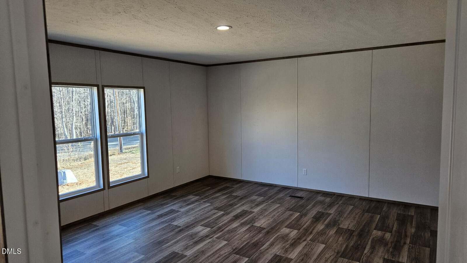 Empty room with white walls, dark wood-style flooring, large windows letting in natural light, and a single ceiling light. A view of trees and outdoors is visible through the windows.