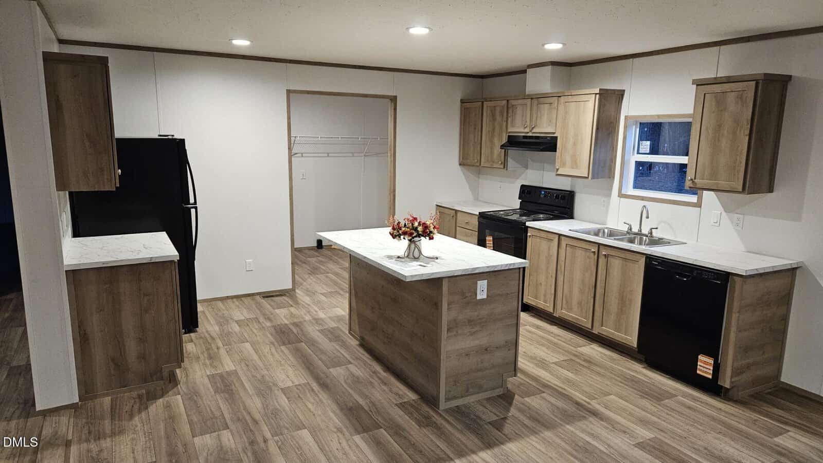 Modern kitchen with wood cabinets, an island with a flower arrangement, black appliances including a stove, refrigerator, and dishwasher, and a walk-in pantry. The flooring is wood-look vinyl. Walls are white.