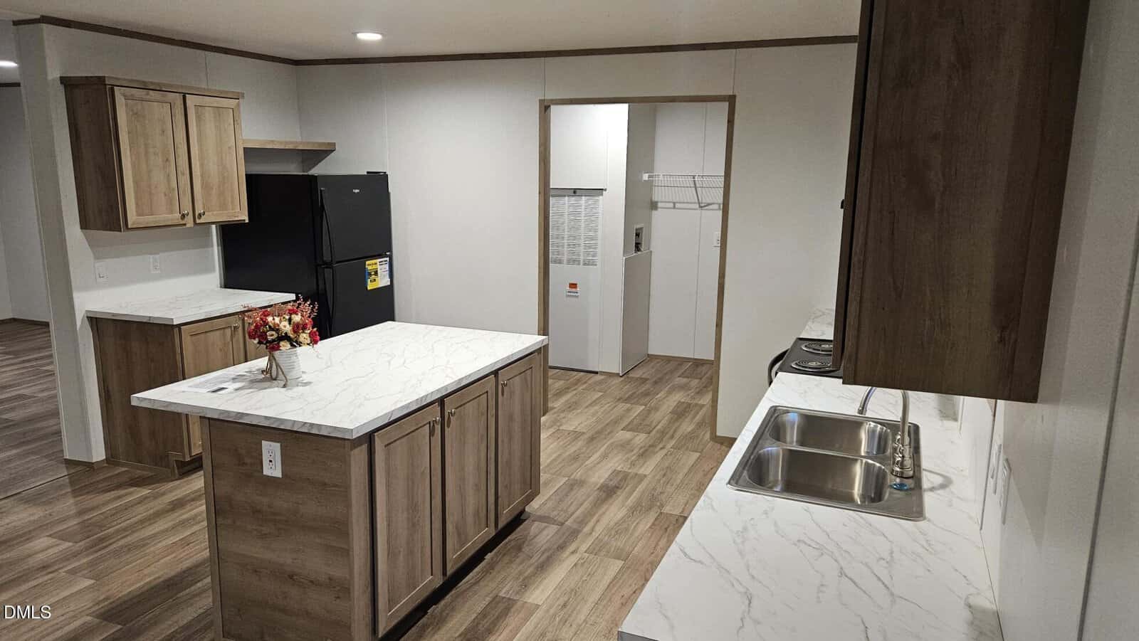 Modern kitchen with wood cabinets, white marble-style countertops, a black refrigerator, and a stainless steel double sink. Adjacent room has a washer and dryer with shelving above. The floor is wood-patterned tile.