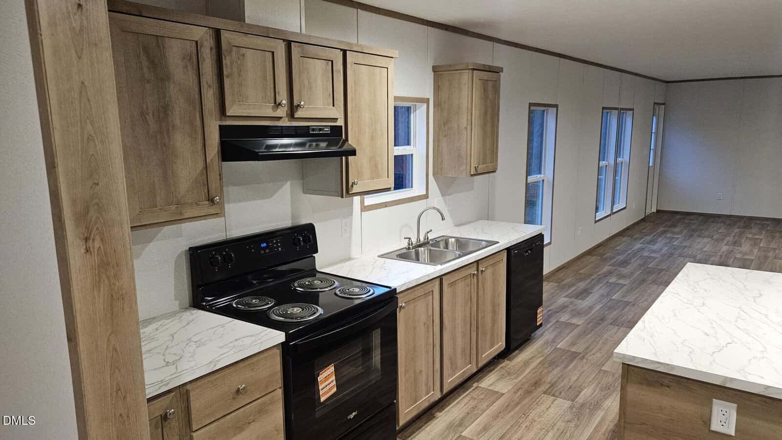 Modern kitchen with light wood cabinets, black appliances, marble-look countertops, double sink, and wood-style flooring. Kitchen opens to a spacious living area with large windows.