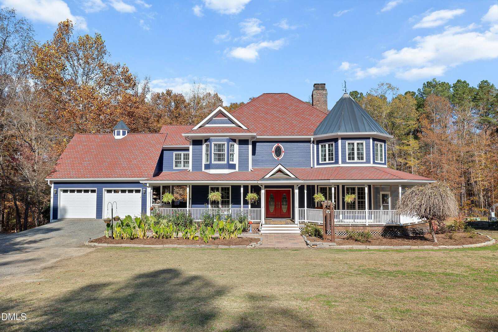 A large two-story blue house with white trim, red roof, and a wraparound porch sits at 1157 Dutchville Drive in Creedmoor, set on a spacious lawn with landscaped garden beds and surrounded by autumn trees under a blue sky.