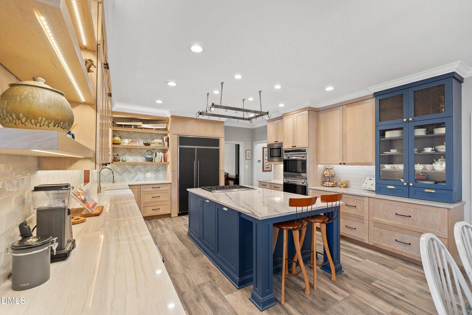 Modern kitchen with light wood cabinets, a navy blue island with marble countertop, stainless steel appliances, open shelves, and two wooden barstools. The floor is wood and the lighting is recessed.
