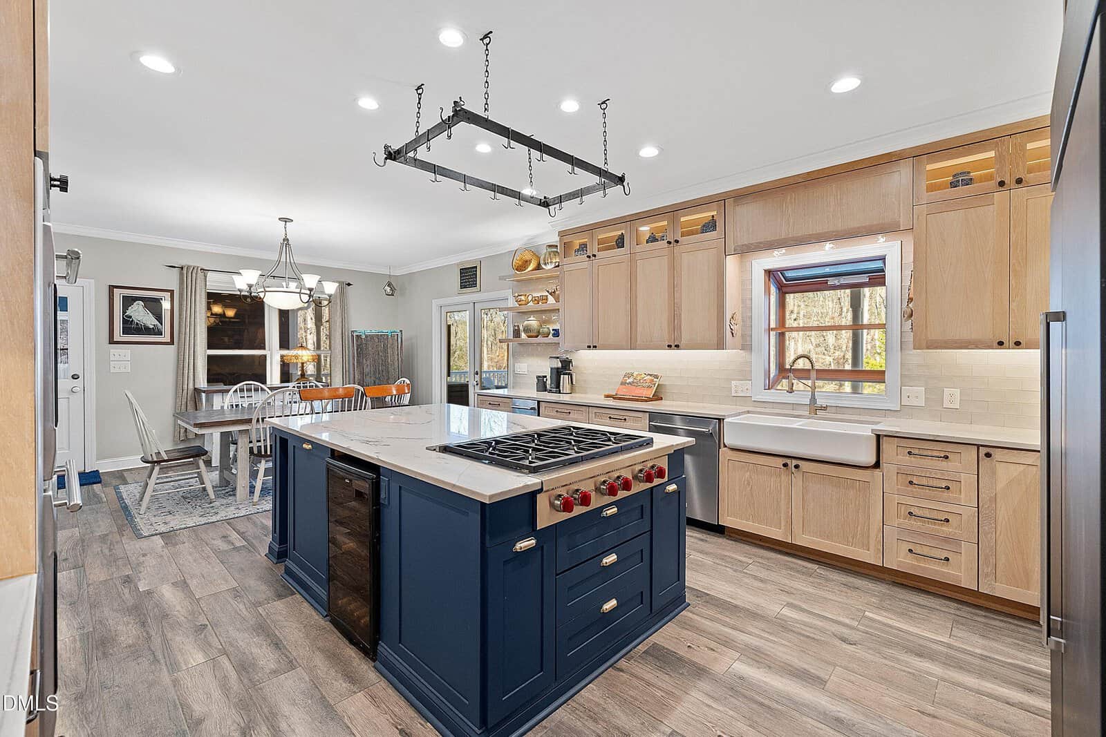 Modern kitchen with wood cabinets, a blue island with stovetop, farmhouse sink, and stainless steel appliances. Dining area with table and chairs is visible in the background near large windows.