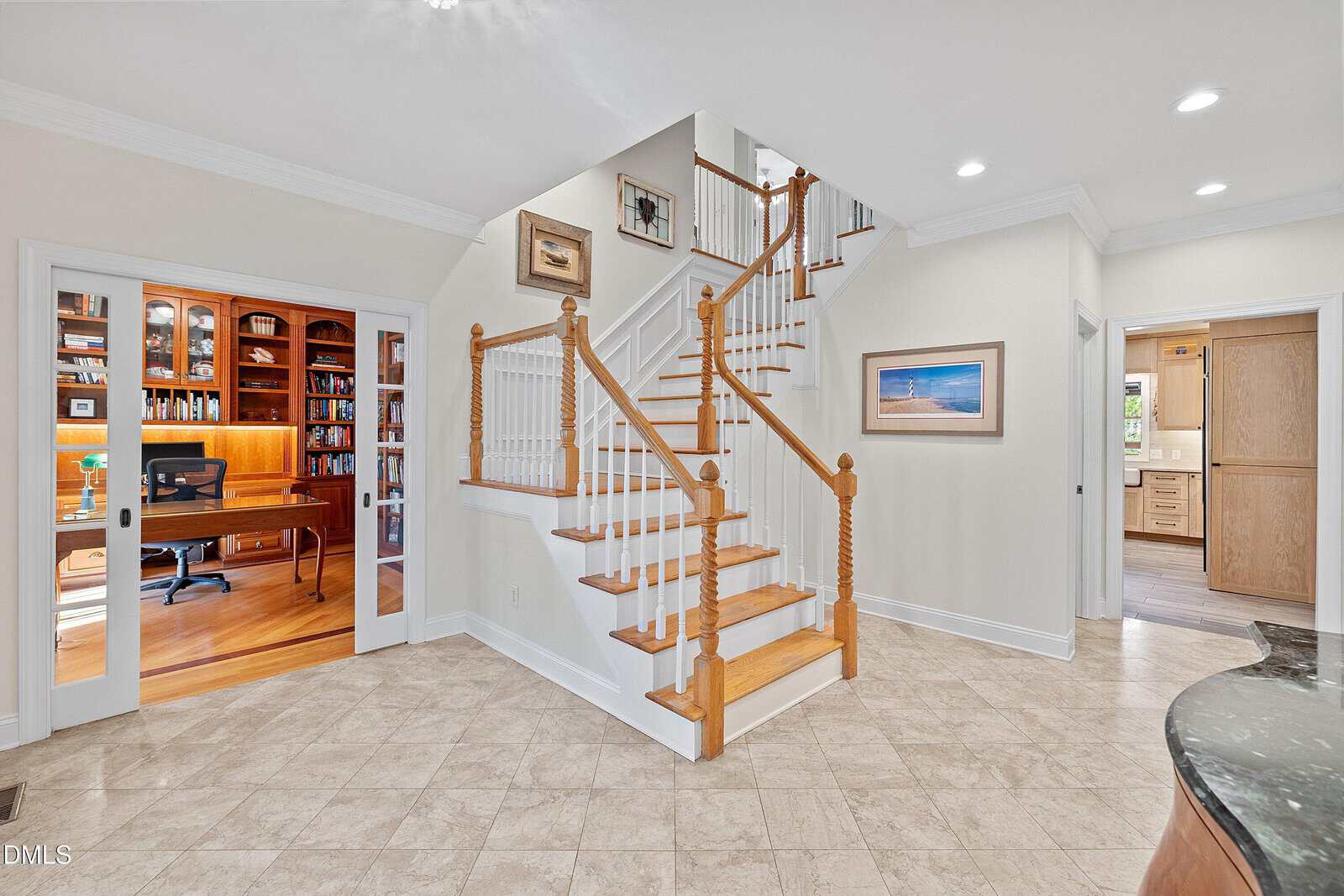 A spacious foyer with beige tile flooring and a wooden staircase. To the left is a home office with built-in bookshelves and a desk. To the right is an open doorway leading to a kitchen area. Framed art hangs on the walls.