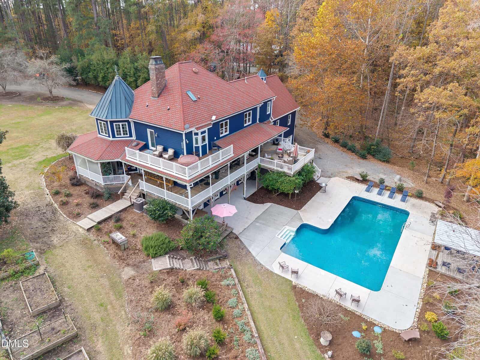 A large two-story blue house with a red roof, wraparound deck, and screened porch overlooks a rectangular swimming pool and patio, surrounded by trees with autumn foliage.