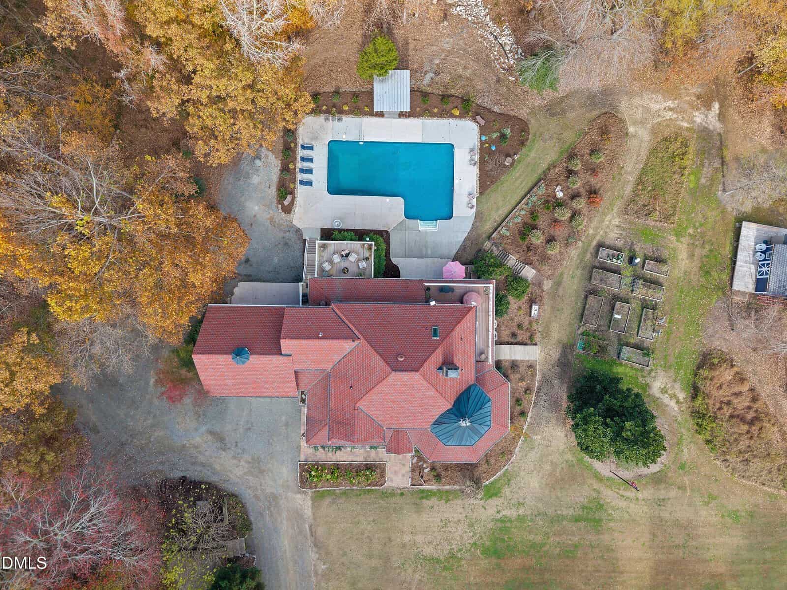 Aerial view of a house with a red roof, a rectangular swimming pool with lounge chairs, a driveway, a small garden with raised beds, and surrounding trees with autumn foliage.