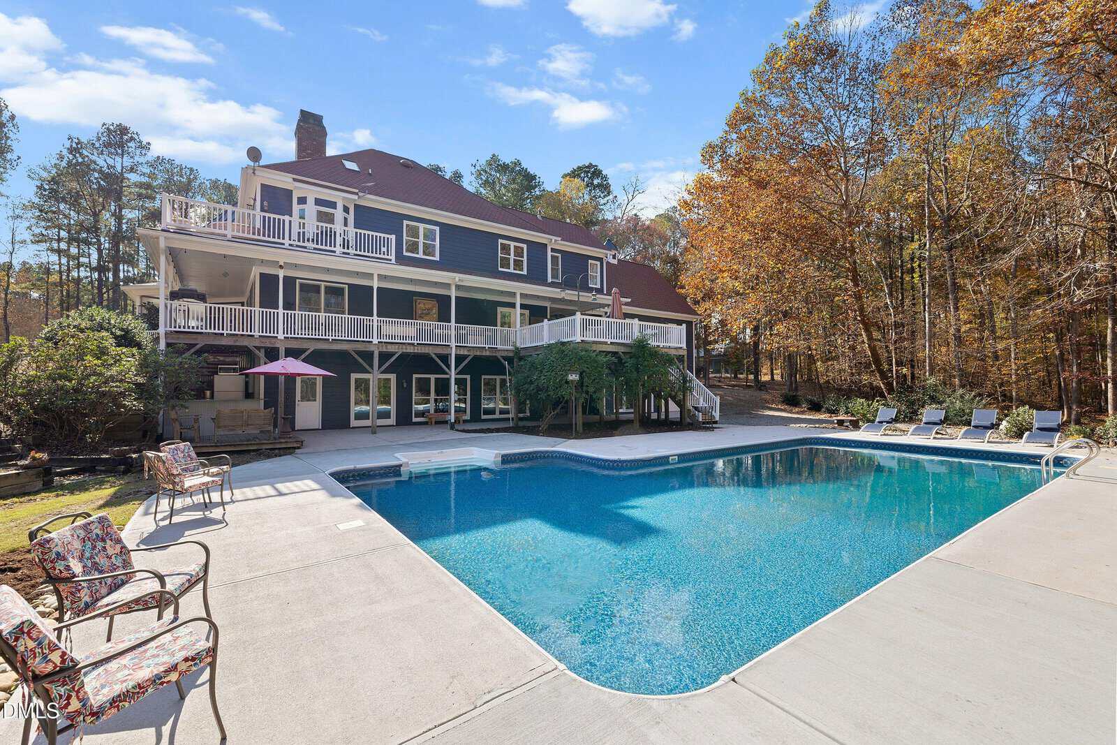A large backyard swimming pool with a patio area featuring lounge chairs, tables, and chairs. A multi-story house with balconies and wooded trees are in the background under a blue sky.