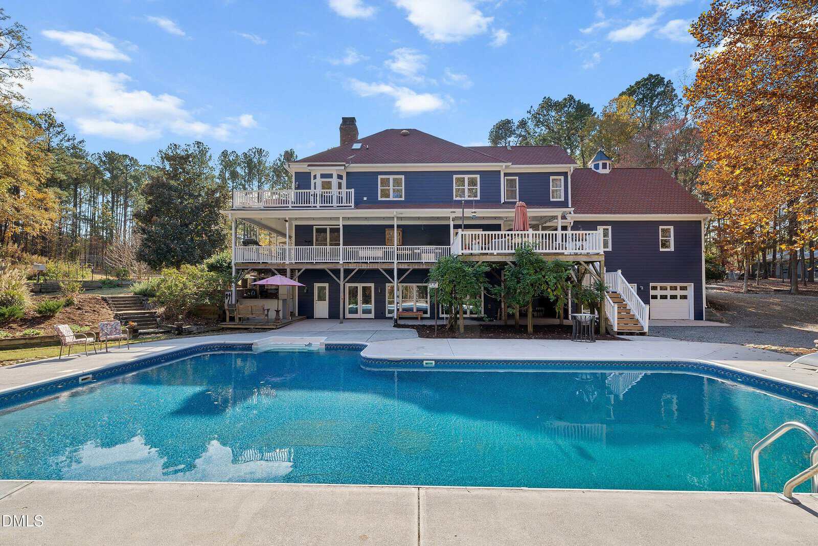 A large house with blue siding, white trim, and red roof sits behind a rectangular swimming pool. The house has multiple balconies, a patio, and is surrounded by trees with autumn foliage.