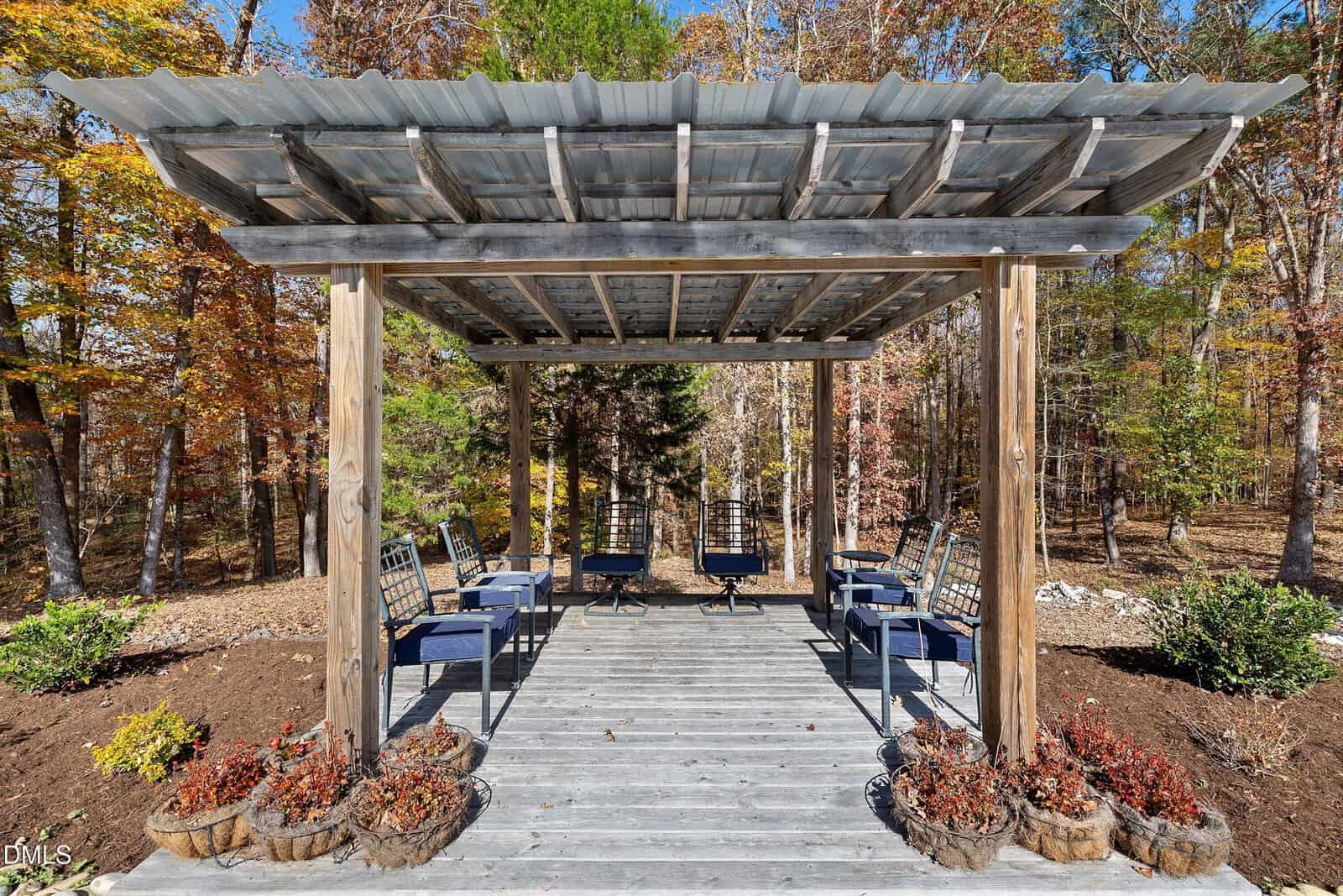 A wooden pergola with a metal roof stands over a deck, surrounded by potted plants and outdoor chairs. Trees with autumn foliage are visible in the background.