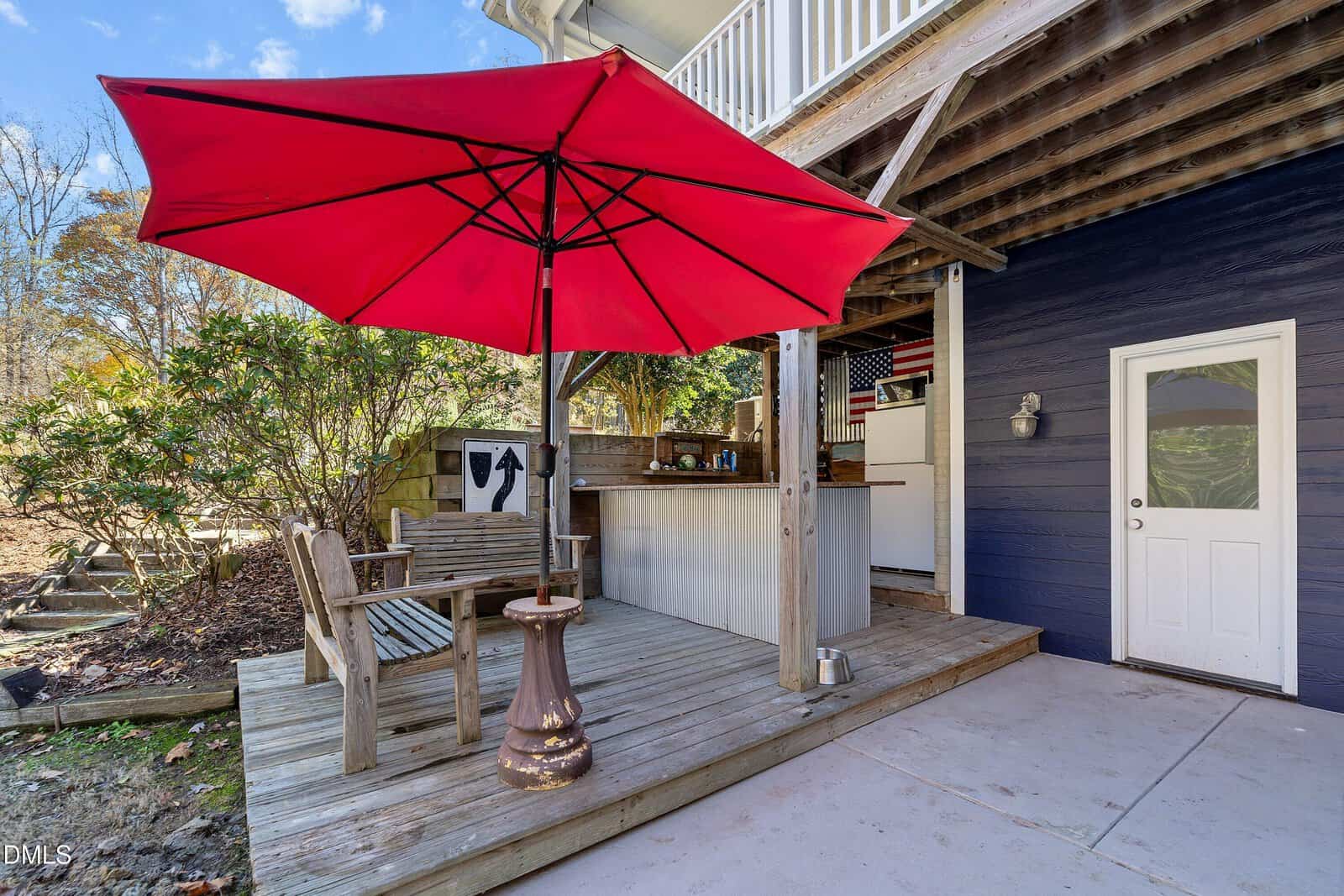 A wooden patio with outdoor seating, a small table, and a large red umbrella. Behind the seating area, there is an outdoor bar under a covered section attached to a dark blue house. Trees and shrubs are in the background.