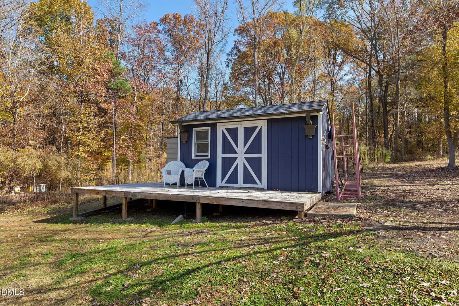 A small blue shed with white trim sits on a wooden deck in a grassy yard, surrounded by autumn trees. Two white chairs and a table are on the deck, and red stairs lead up the hill on the right.