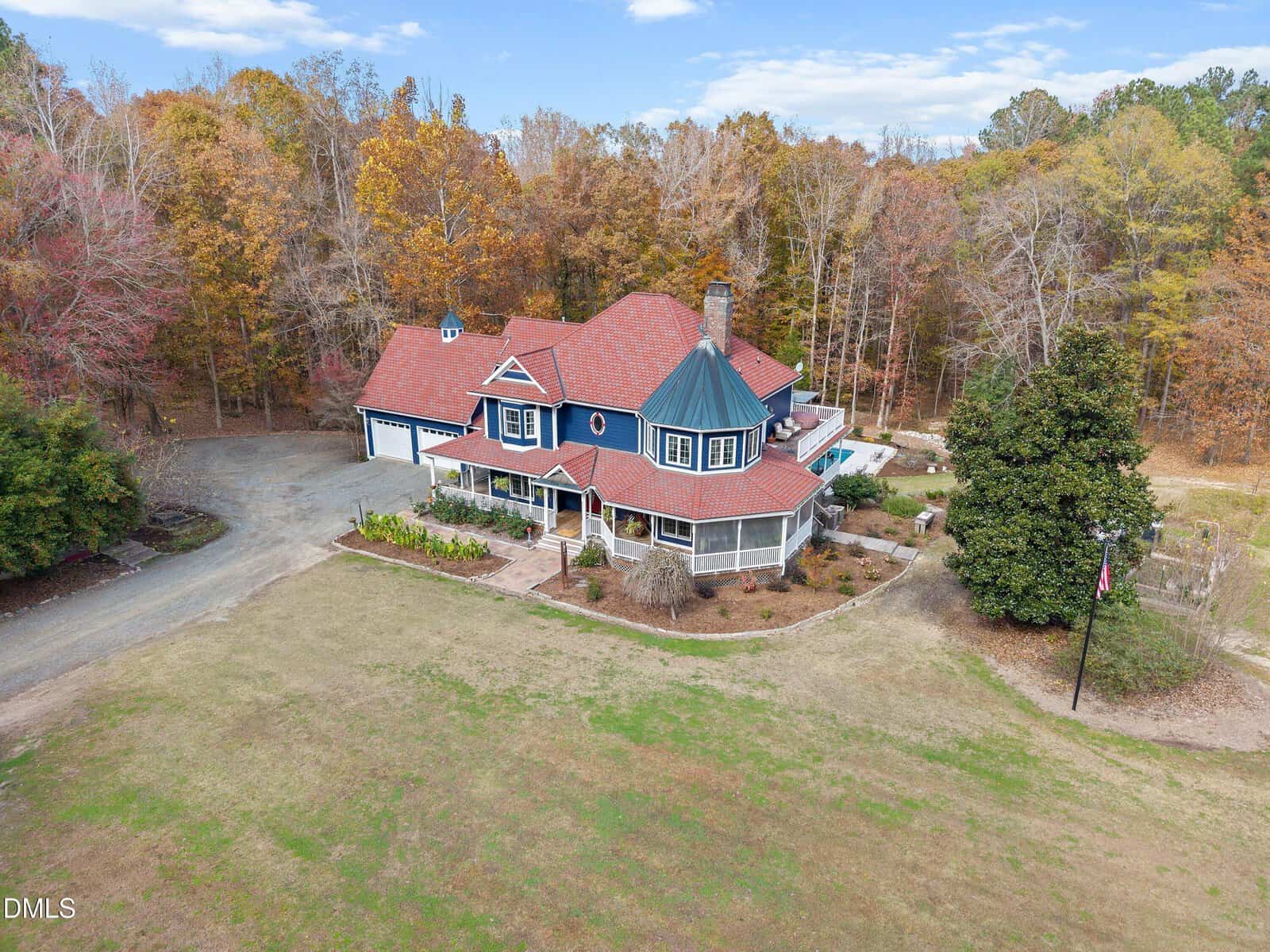 A large two-story house with red roofs and blue siding sits surrounded by trees with autumn foliage. The property has a circular driveway, attached garage, and landscaped yard. The sky is partly cloudy.