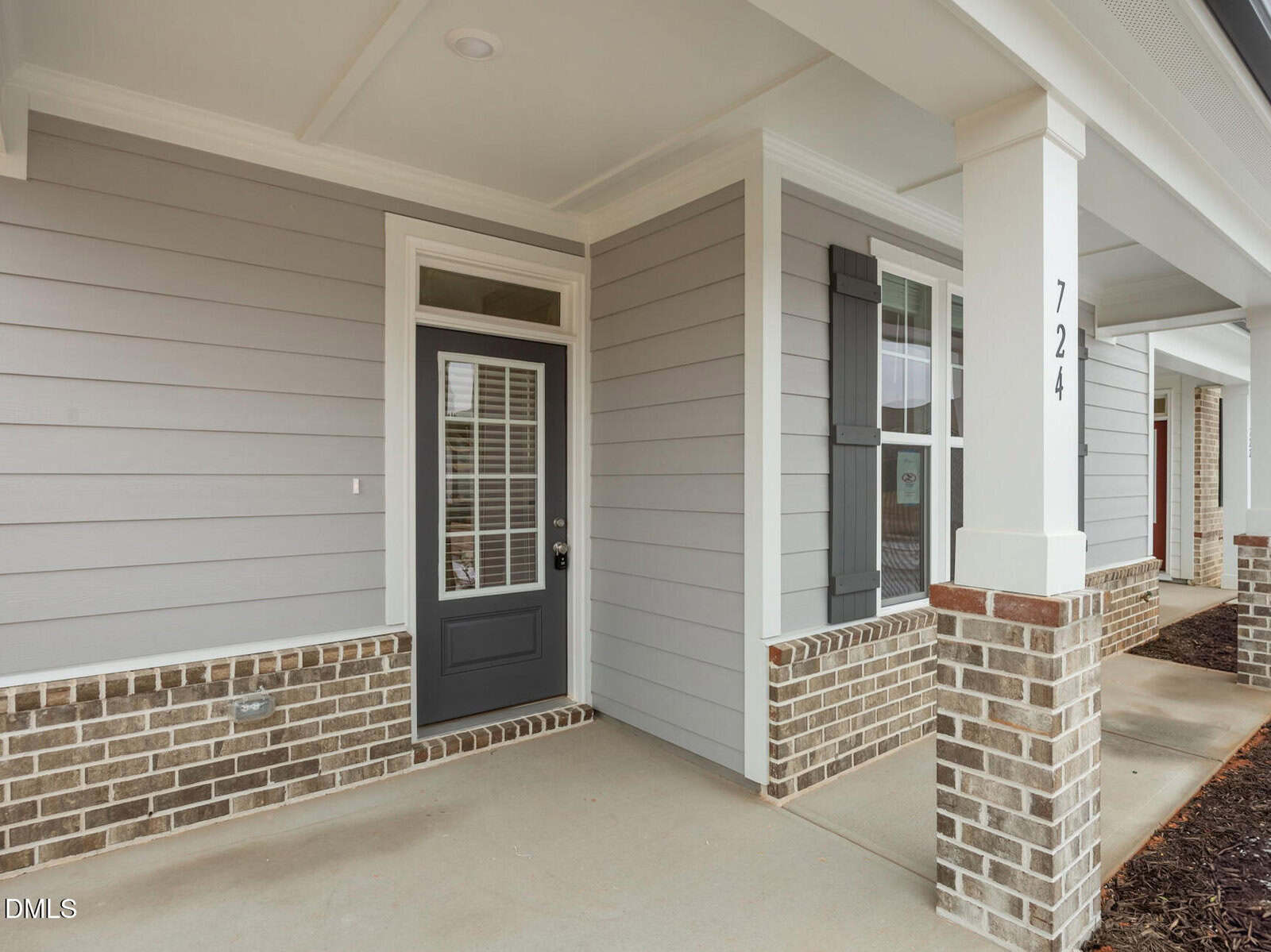 A covered front porch with light gray siding, white trim, and brick accents. A glass-paneled gray front door is next to a window with dark shutters. The house number 724 is visible on a white column.