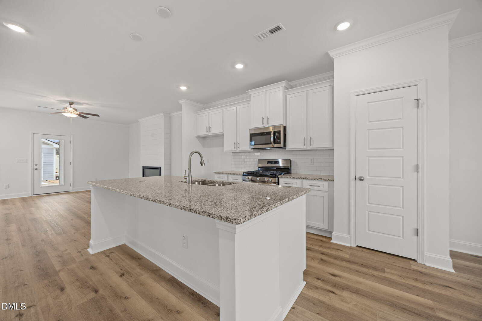 Modern kitchen with white cabinets, stainless steel appliances, granite countertops, and a large island. The space has wood flooring, recessed lighting, and an open layout leading to another room with a ceiling fan.