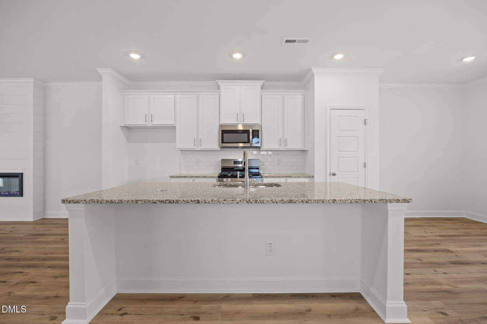 Modern kitchen with white cabinets, granite countertops, stainless steel appliances, and a large island. The room has wood flooring, recessed lighting, and a minimalist, clean design.