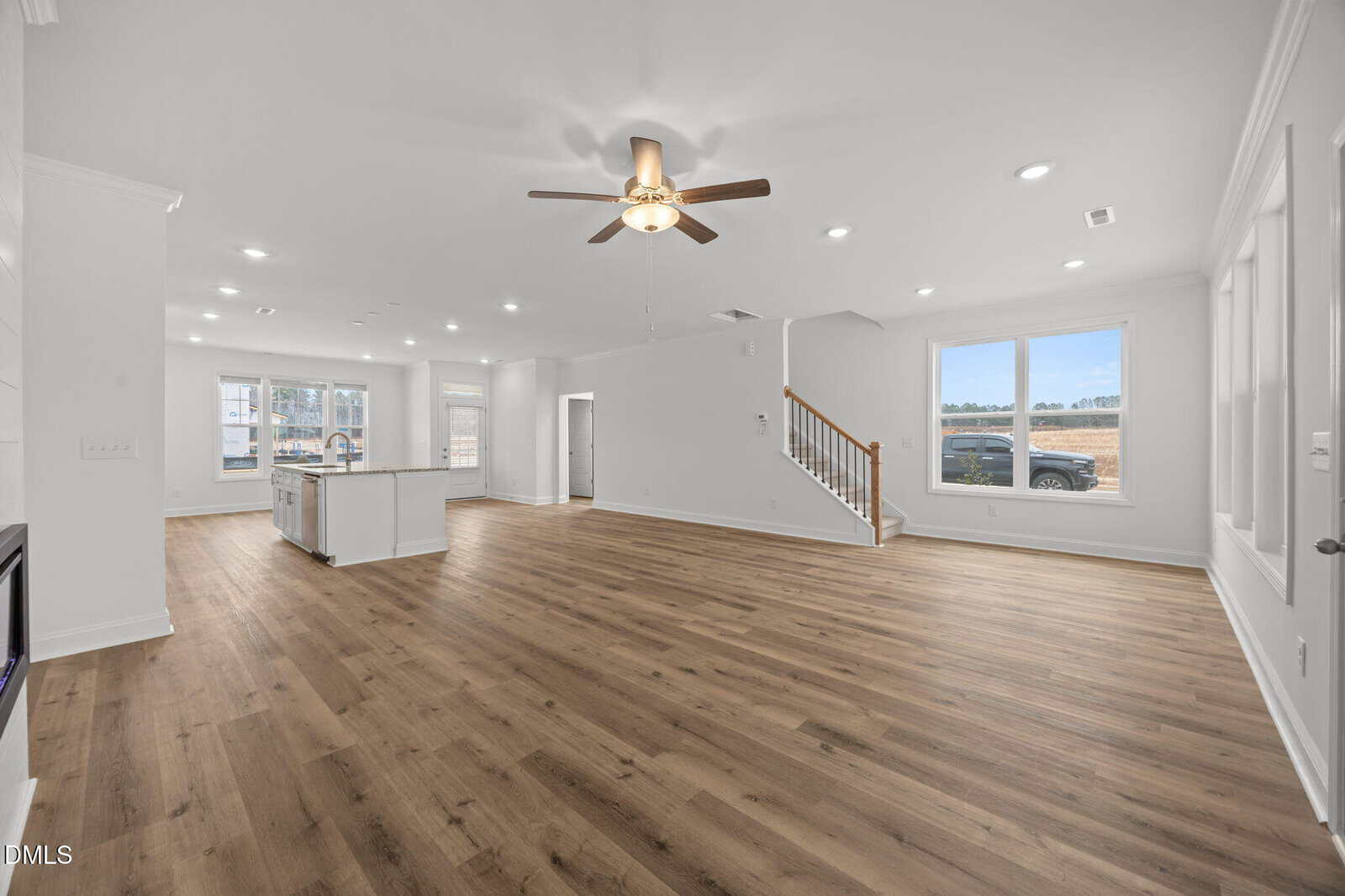 Spacious, modern open-plan living area with light wood floors, white walls, ceiling fan, large windows, and a staircase leading upstairs. Kitchen with island visible in the background. Natural light fills the room.