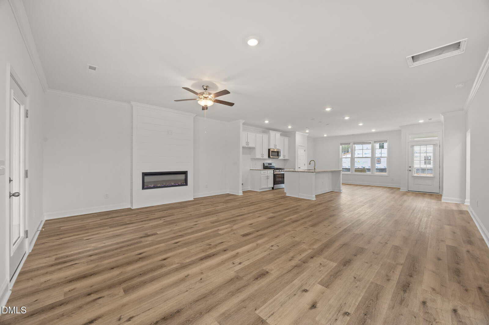 Open-concept living space with wood floors, white walls, modern kitchen with island, electric fireplace, ceiling fan, and large windows letting in natural light.