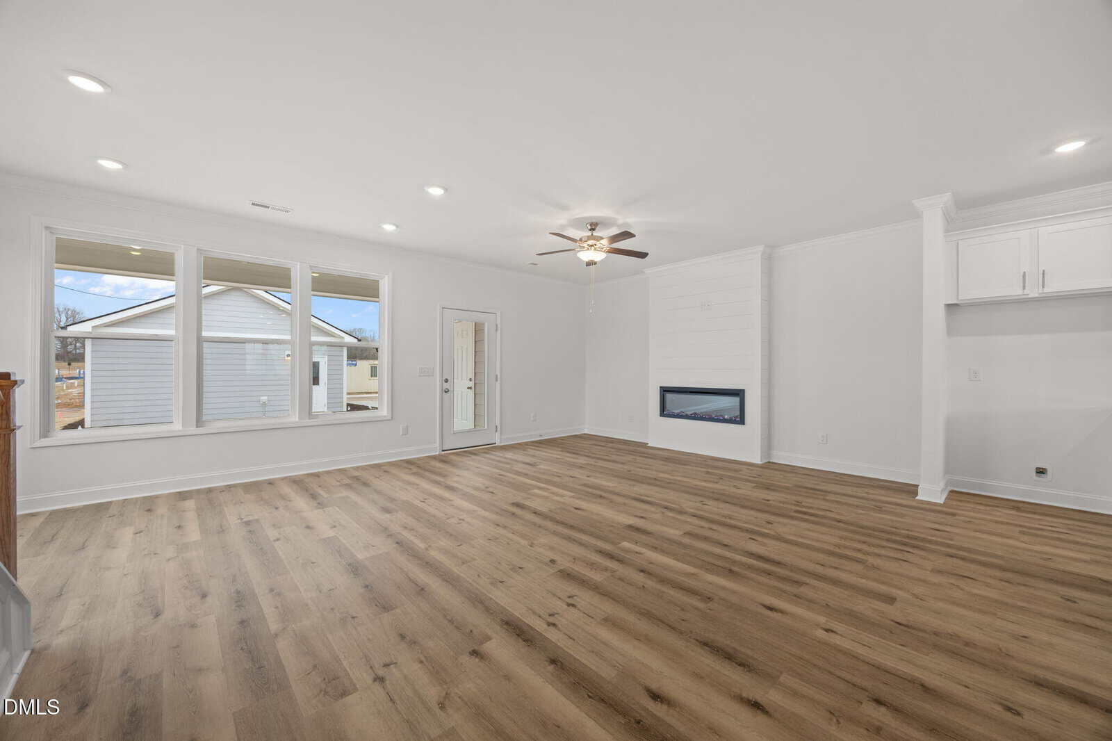 Spacious, empty living room with light wood flooring, white walls, large windows, a ceiling fan, a modern built-in fireplace, and a door leading outside. Natural light fills the open, clean space.