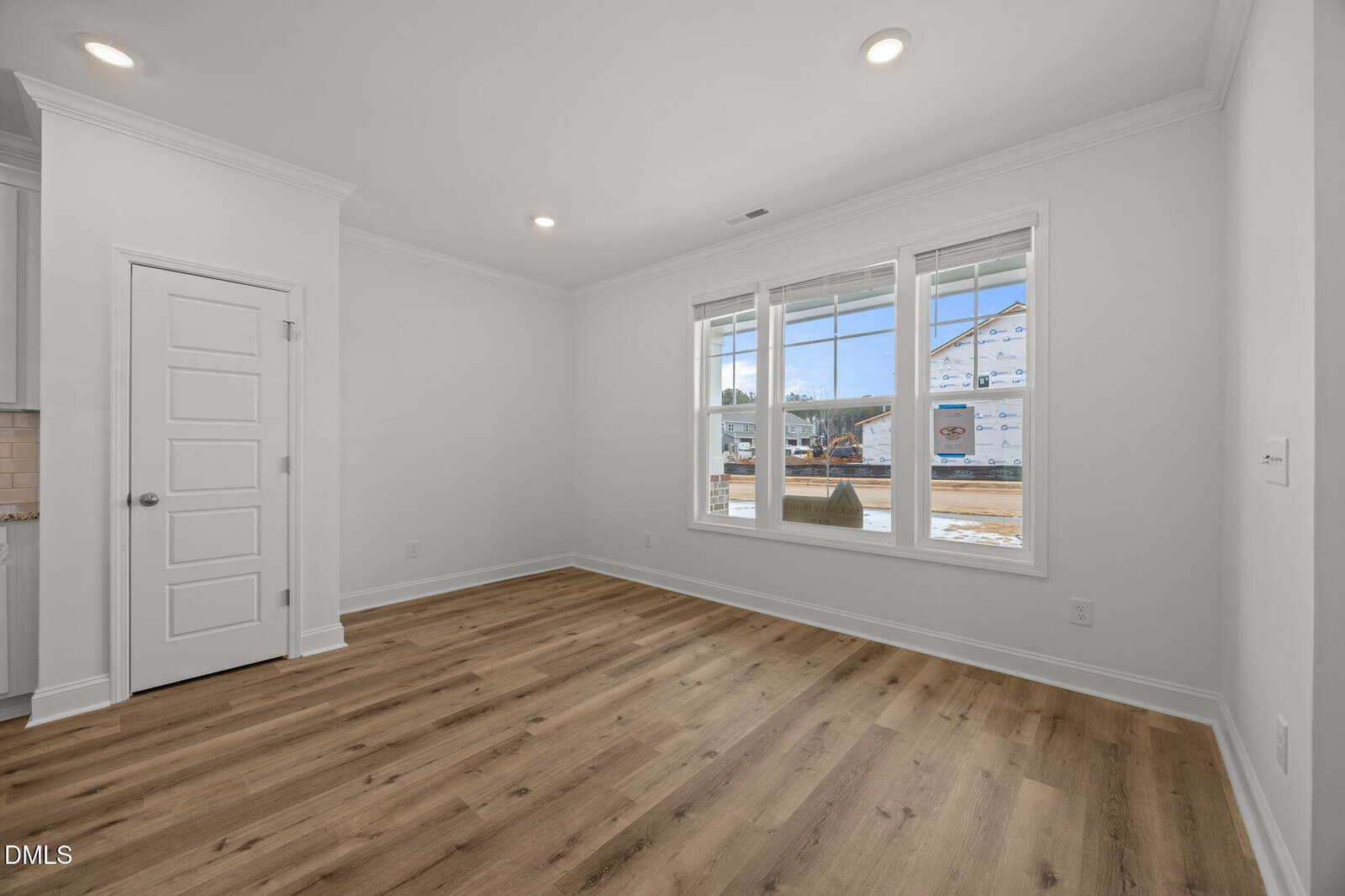 A bright, empty room with white walls, wide windows letting in natural light, recessed ceiling lights, wood-style flooring, and a white door; construction is visible outside the window.