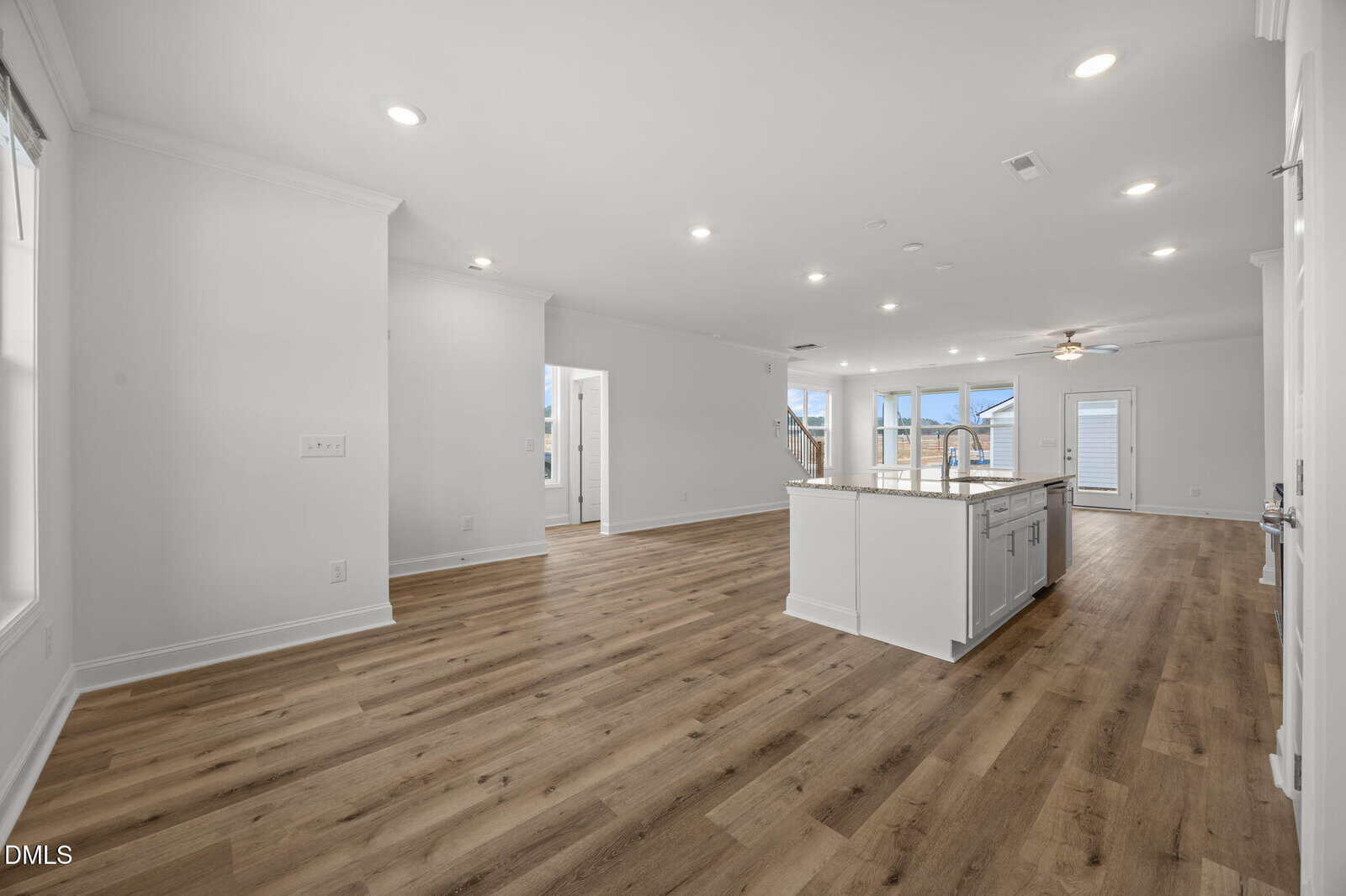 Spacious modern open-concept living area with wood flooring, white walls, recessed lighting, and a central kitchen island. Large windows bring in natural light, and a ceiling fan is visible near the back door.