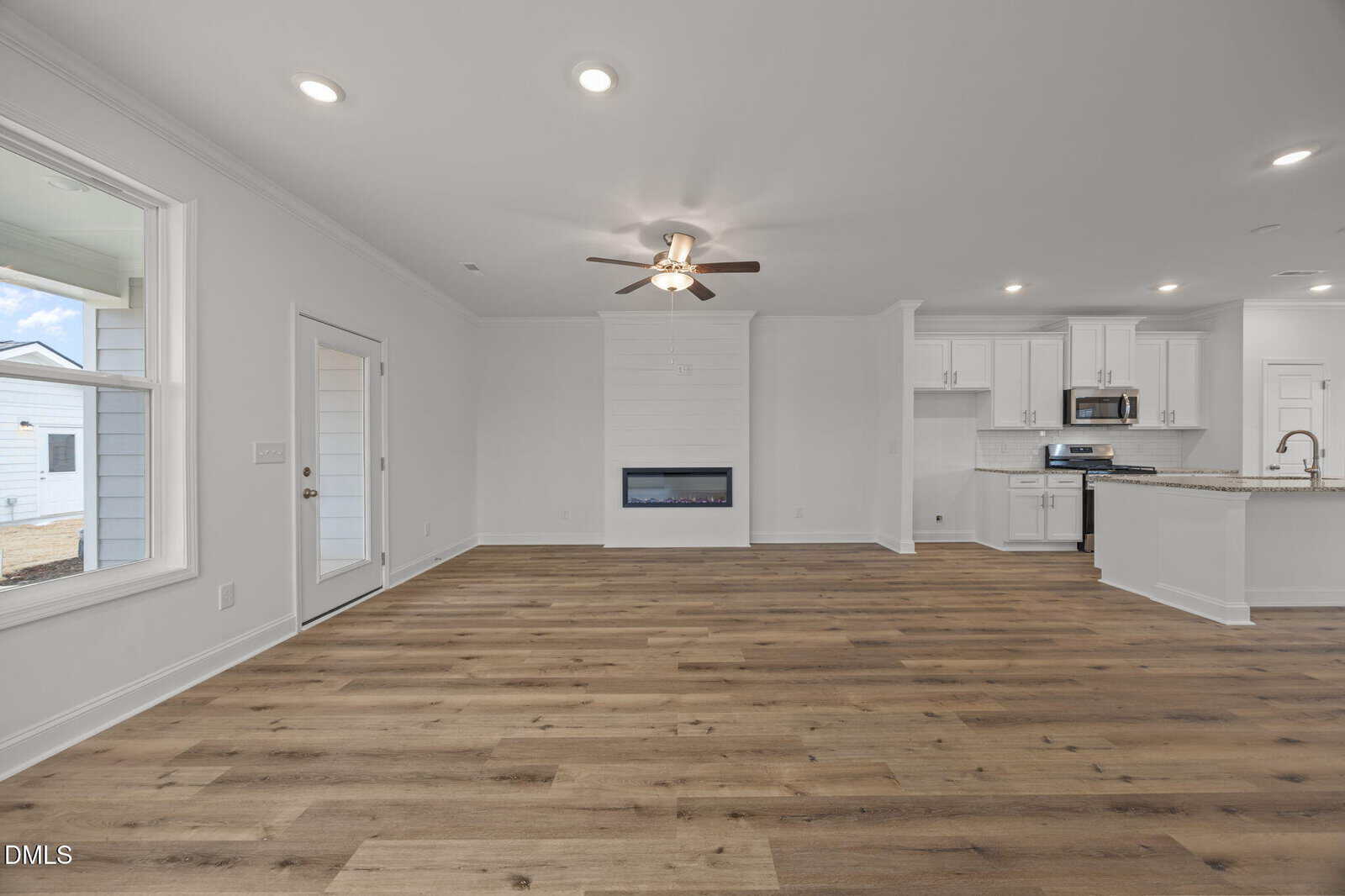 Open-concept living area with light wood flooring, white walls, ceiling fan, built-in fireplace, large windows, and a modern kitchen with white cabinets, granite countertops, and stainless steel appliances.