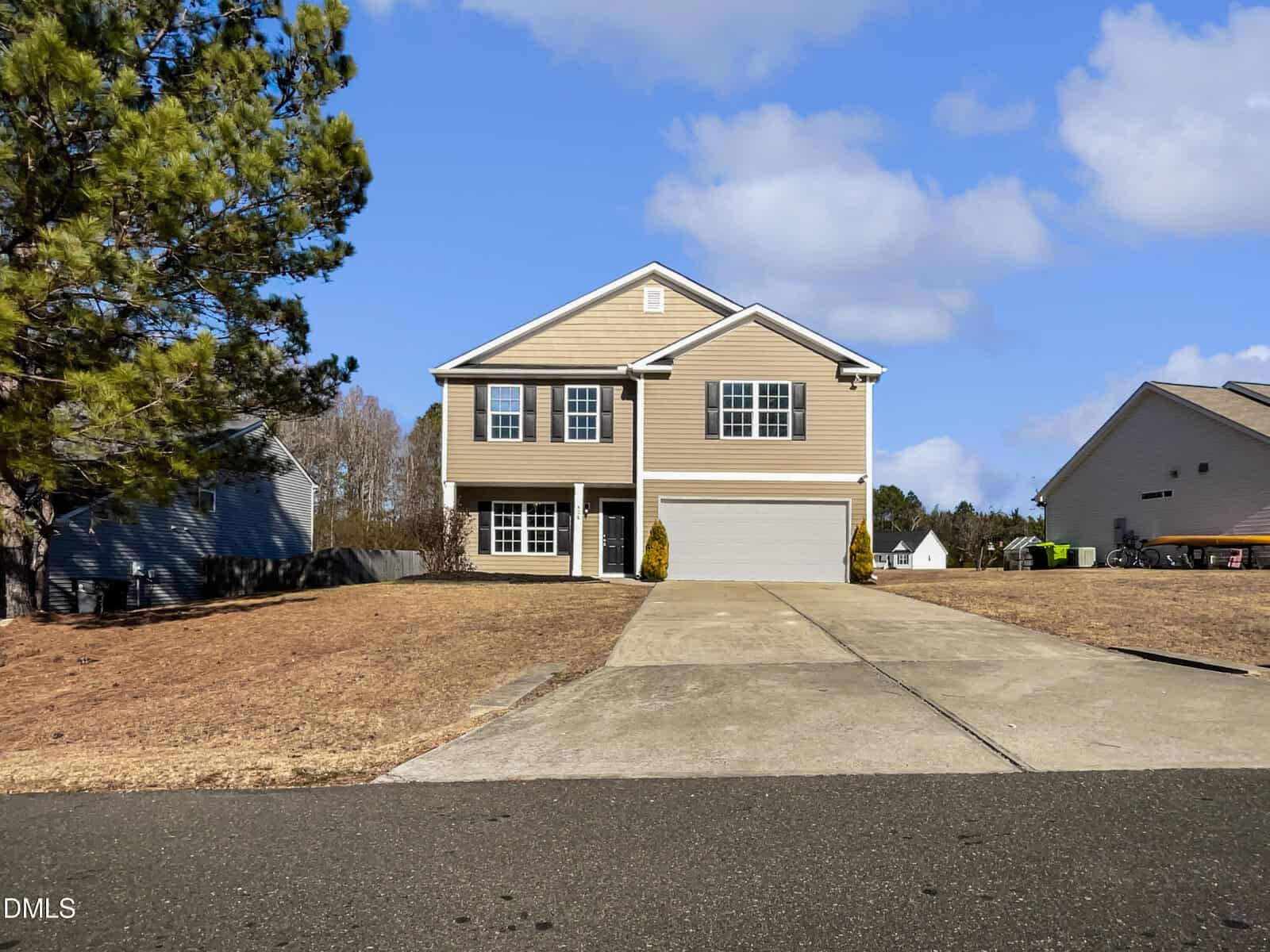 A two-story beige house with white trim and a double garage sits at the end of a wide driveway at 506 Wild Goose Lane, surrounded by dry brown grass and neighboring homes under a blue sky with scattered clouds.