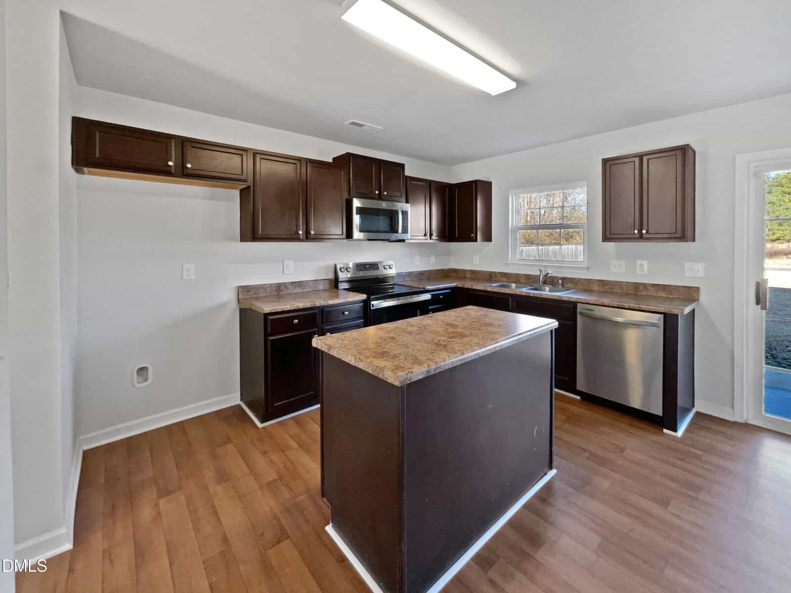 A modern kitchen with dark brown cabinets, stainless steel appliances, a center island, wood flooring, and a window above the sink offering outside light. The room is clean and empty.