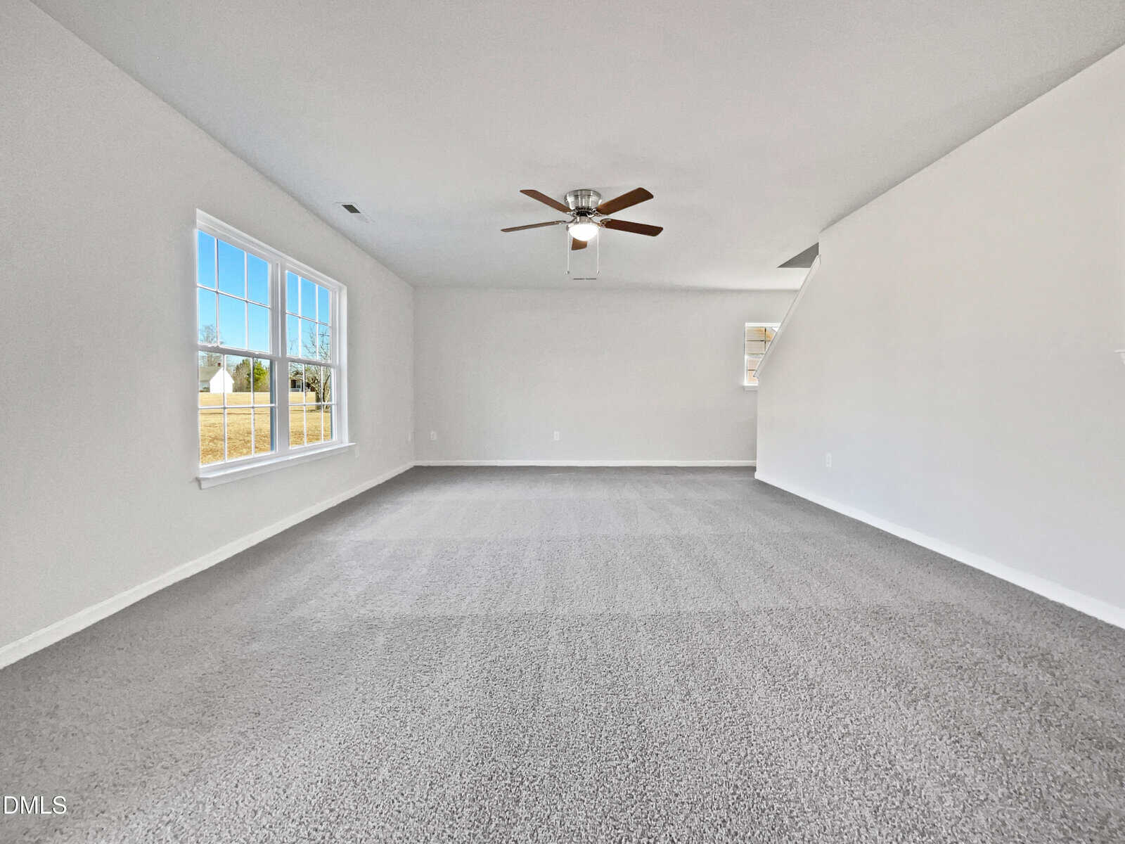 A spacious, empty room with light gray carpet, white walls, a ceiling fan with a light fixture, a large window on the left, and a small window near a stairway on the right.