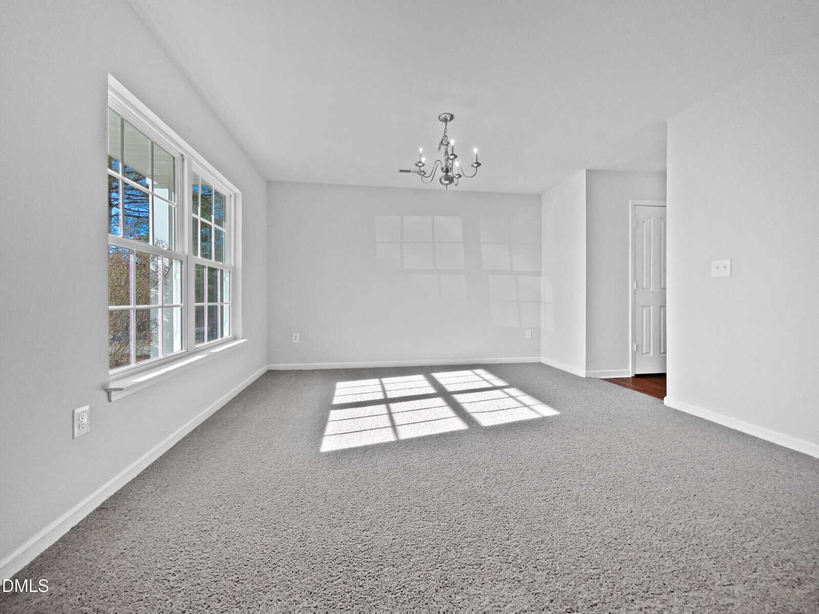 A bright, empty room with light gray carpet, white walls, a large window on the left letting in natural light, a white ceiling, a simple chandelier, and a closed white door in the back right corner.